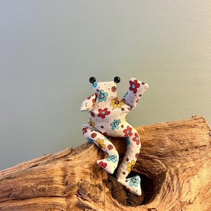 Könnte beinhalten: Ein Stofffrosch aus weißem Stoff mit einem bunten Blumenmuster aus roten, gelben und hellblauen Blüten. Der Frosch hat zwei kleine schwarze Perlenaugen auf dem Kopf und ist mit angewinkelten Gliedmaßen in einer sitzenden Position dargestellt.