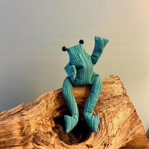 Könnte beinhalten: Ein Stofffrosch aus blaugrünem Stoff mit dünnen weißen vertikalen Streifen. Der Frosch hat zwei kleine schwarze Perlenaugen, lange baumelnde Beine und einen erhobenen Arm. Das Material weist eine sichtbare Textur auf und die Figur sitzt aufrecht.