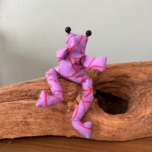 Könnte beinhalten: Ein Stofffrosch in Hellviolett mit einem roten Linienmuster. Der Frosch hat zwei schwarze, kugelförmige Augen auf dem Kopf, lange Gliedmaßen und einen flexiblen Körper, der so gestaltet ist, dass er aufrecht auf einer Oberfläche sitzen kann.