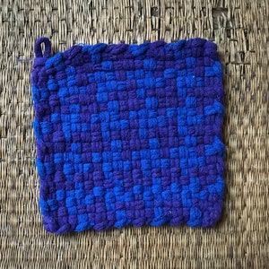 Op de afbeelding: Een vierkante, geweven pannenlap in blauwe en paarse tinten. De pannenlap heeft een lus om op te hangen en is gemaakt van dik, getextureerd garen. Het weefpatroon creëert een dambordeffect met de twee kleuren.