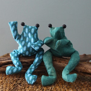 Könnte beinhalten: Zwei handgefertigte Frosch-Plüschtiere. Ein Frosch ist türkis mit einem blau-weißen Muster, der andere ist einfarbig türkis. Beide haben schwarze Knopfaugen und sitzen auf einem Stück Treibholz. Die Frösche befinden sich auf einer gewebten Oberfläche.