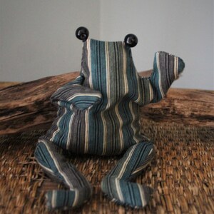 Könnte beinhalten: Ein handgefertigtes Frosch-Plüschtier mit einem gestreiften Muster in Türkis, Grau und Weiß. Der Frosch hat große schwarze Augen und ist mit einem erhobenen Arm posiert. Das Spielzeug sitzt auf einer gewebten Oberfläche.