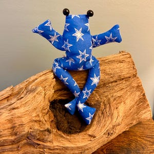 Könnte beinhalten: Ein Stofffrosch aus blauem Stoff mit weißem Sternenmuster. Der Frosch hat zwei schwarze Perlenaugen, ausgestreckte Arme und lange Beine. Er sitzt in einer aufrechten Position mit gekreuzten Beinen und weist eine einfache, weiche Konstruktion auf.