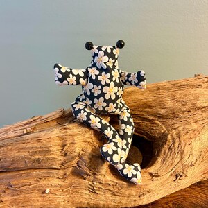 Könnte beinhalten: Ein Stofffrosch aus schwarzem Stoff mit einem weißen und gelben Gänseblümchenmuster. Der Frosch hat zwei kleine schwarze Perlenaugen auf dem Kopf, ausgestreckte Arme und lange Beine, die in einer sitzenden Position angeordnet sind.