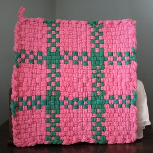 May include: A square, woven hot pad in a vibrant pink color with a green cross pattern. The hot pad is made of thick, textured material and has a loop for hanging. The design is a simple, yet eye-catching, geometric pattern.