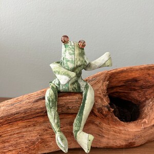 Könnte beinhalten: Eine handgefertigte Stofffroschpuppe mit grün-weiß gemustertem Stoff, die auf einem Stück Treibholz sitzt. Der Frosch hat braune Perlenaugen und ist mit einem erhobenen Arm posiert. Das Treibholz hat ein natürliches, verwittertes Aussehen.