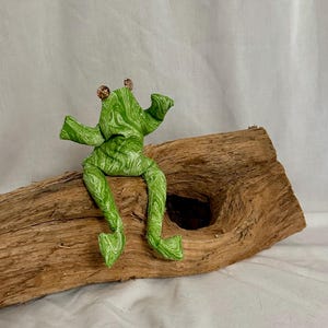 Könnte beinhalten: Eine kleine Stofffrosch-Figur aus grün-weiß marmoriertem Stoff. Der Frosch hat zwei kleine, runde, hellbraune Perlenaugen auf dem Kopf. Die Arme sind erhoben und die Beine hängen in einer sitzenden Position herab.