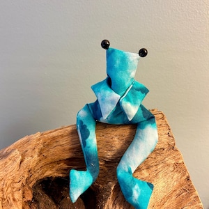 Könnte beinhalten: Ein Stofffrosch mit einem Batikmuster in leuchtenden Blau- und Weißtönen. Der Frosch hat zwei kleine schwarze Perlenaugen oben am Kopf und lange, herabhängende Beine, die sich vom Körper aus nach unten erstrecken.