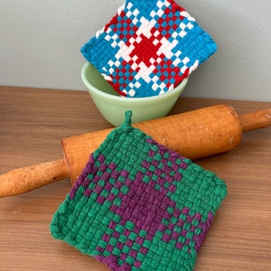 May include: Two woven potholders in the image. One is blue with a red and white pattern, and the other is green with a purple pattern. A light green bowl and a wooden rolling pin are also in the image. The potholders are handmade.