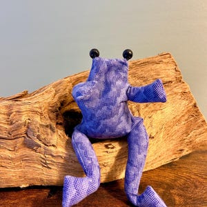 Könnte beinhalten: Ein Stofffrosch aus blauem Stoff mit einem dunkleren blauen, gesprenkelten Muster. Der Frosch hat zwei kleine schwarze Perlenaugen auf dem Kopf, zwei zur Seite ausgestreckte Arme und zwei lange, an den Knien angewinkelte Beine in Sitzposition.