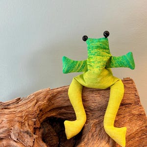 Könnte beinhalten: Ein Stofffrosch aus Textil mit einem gesprenkelten grün-gelben Muster. Der Frosch hat zwei schwarze Perlenaugen, ausgestreckte Arme und lange, herabhängende Beine. Der Körper besteht aus einem strukturierten, lindgrünen Stoffmaterial.