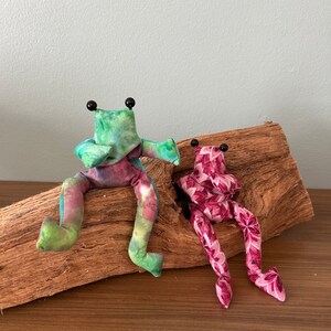 Könnte beinhalten: Zwei Stofffroschpuppen mit schwarzen Perlenaugen und ausgestreckten Armen. Ein Frosch ist im Batikstil in Grün-, Blau- und Rosatönen gehalten. Der andere Frosch ist mit einem rosa Blumenmuster versehen. Beide Frösche sitzen auf einem Stück Treibholz.