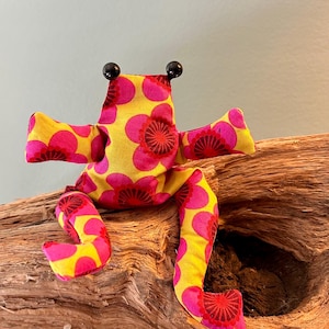 Könnte beinhalten: Ein Stofffrosch aus gelbem Stoff mit einem sich wiederholenden Muster aus rosa und dunkelroten Blumen. Der Frosch hat zwei schwarze Perlenaugen auf dem Kopf, zwei ausgestreckte Arme und zwei lange Beine, die nach unten ragen.