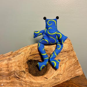 Könnte beinhalten: Ein Stofffrosch aus blauem Stoff mit gelbem Wirbelmuster. Der Frosch hat zwei kleine schwarze Perlenaugen auf dem Kopf, lange Gliedmaßen und sitzt aufrecht, wobei die Beine in einer entspannten, sitzenden Position nach unten hängen.