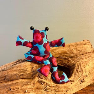 Könnte beinhalten: Ein Stofftier in Froschform mit hellblauem Hintergrund und einem sich wiederholenden Muster aus dunkelvioletten und leuchtend pinken kreisförmigen Blumenmotiven. Das Spielzeug hat zwei kleine schwarze, kugelförmige Perlen als Augen.