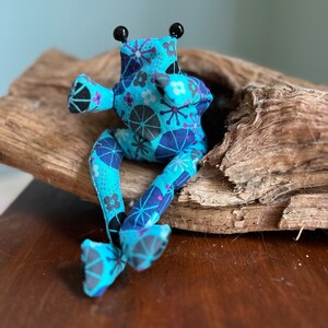 Könnte beinhalten: Eine handgefertigte Stofffroschpuppe mit blauem Blumenmuster. Der Frosch hat schwarze Perlenaugen und sitzt auf einem Stück Treibholz. Die Arme und Beine der Puppe sind ausgestreckt, was ein skurriles und charmantes Design ergibt.