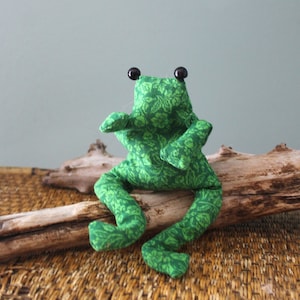 Könnte beinhalten: Ein Stofffrosch aus grünem Stoff mit einem sich wiederholenden Blumen- und Blattmuster. Die Plüschfigur hat zwei kleine, runde, schwarze Perlenaugen auf dem Kopf sowie lange, herabhängende Beine und zwei nach vorne ausgestreckte Arme.