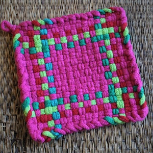 Op de afbeelding: Een vierkante, geweven pannenlap in felroze, met accenten van rood, groen en teal. De randen zijn afgewerkt met een gevlochten bies. Er is een lus bevestigd om op te hangen. De pannenlap is ontworpen voor gebruik in de keuken.