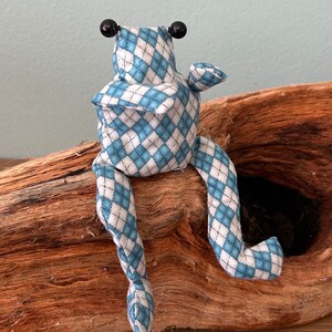 Könnte beinhalten: Eine handgefertigte Stofffroschpuppe mit blauem und weißem Rautenmuster. Der Frosch hat schwarze Perlenaugen und sitzt auf einem Stück Treibholz. Die Arme und Beine des Frosches sind ausgestreckt.