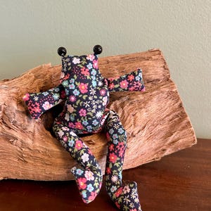 Könnte beinhalten: Eine handgefertigte Stofffroschpuppe mit einem Blumenmuster in Rosa, Blau und Weiß auf marineblauem Hintergrund. Der Frosch hat schwarze Perlenaugen und sitzt auf einem Stück Treibholz. Die Arme und Beine der Puppe sind ausgestreckt.