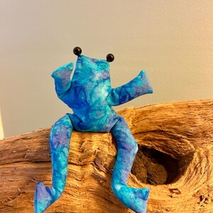 Könnte beinhalten: Ein Stofffrosch aus gepolstertem Material mit einem marmorierten Muster in Blau- und Violetttönen. Der Frosch hat zwei kleine schwarze Perlenaugen auf dem Kopf, ausgestreckte Arme und lange, angewinkelte Beine, die vom Körper herabhängen.