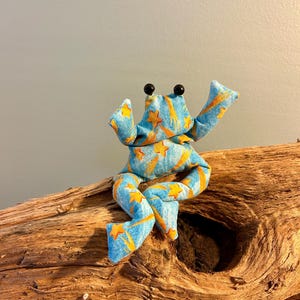 Könnte beinhalten: Ein Stofffrosch-Spielzeug mit einem hellblauen Grundton und einem wiederkehrenden Muster aus orangefarbenen Sternschnuppen. Der Plüschfrosch hat zwei kleine, runde, schwarze Kunststoffaugen und sitzt in einer Position mit ausgestreckten Gliedmaßen.
