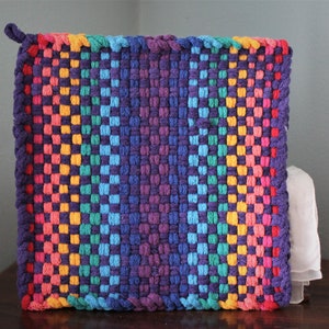 May include: A square, woven hot pad in a vibrant rainbow of colours. The design features a gradient from red to purple, with a loop for hanging. The woven texture is visible, showcasing the craftsmanship of the handmade item.