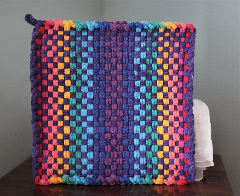 May include: A square, woven hot pad in a vibrant rainbow of colours. The design features a gradient from red to purple, with a loop for hanging. The woven texture is visible, showcasing the craftsmanship of the handmade item.