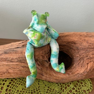 Könnte beinhalten: Eine handgefertigte Stofffroschpuppe mit grünem und blauem Batikmuster. Der Frosch hat grüne Edelsteinaugen und sitzt auf einem Stück Treibholz. Eine grüne gehäkelte Tischdecke befindet sich im Vordergrund.
