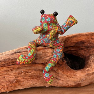 Könnte beinhalten: Eine farbenfrohe Stofffroschpuppe mit schwarzen Perlenaugen und ausgestreckten Armen. Der Frosch ist aus gemustertem Stoff in Rot, Gelb, Blau und Grün gefertigt. Er sitzt auf einem Stück Treibholz.