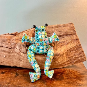 Könnte beinhalten: Ein Stofffrosch aus hellblauem Stoff mit einem bunten Blumenmuster aus kleinen Kreisen in Rosa, Gelb, Grün und Weiß. Der Frosch hat zwei kleine schwarze Perlenaugen auf dem Kopf und lange Gliedmaßen, die in einer sitzenden Position sind.