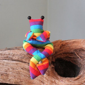 May include: A colorful, handmade stuffed toy frog with a rainbow striped pattern. The frog has black button eyes and is posed on a piece of driftwood. The toy's vibrant colors include red, orange, yellow, green, blue, and purple.