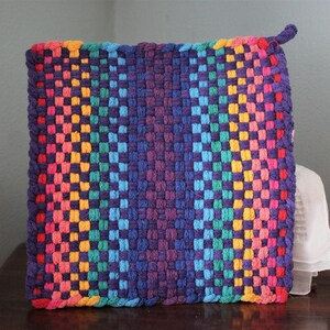 May include: A square, woven hot pad with a rainbow gradient design. The colours transition from pink, orange, yellow, green, and blue to a deep purple in the centre. A small loop is attached to one corner for hanging.