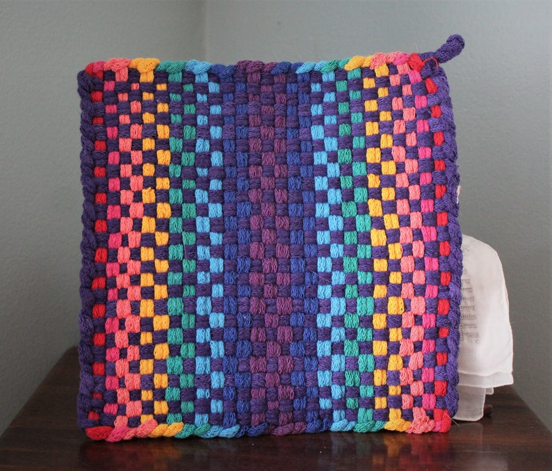 May include: A square, woven hot pad with a rainbow gradient design. The colours transition from pink, orange, yellow, green, and blue to a deep purple in the centre. A small loop is attached to one corner for hanging.