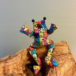 Könnte beinhalten: Ein Stofffrosch aus Textil mit einem bunten Puzzleteilmuster in Rot, Blau, Gelb, Grün und Violett. Der Frosch hat zwei kleine schwarze Perlenaugen auf dem Kopf und ist mit ausgestreckten Armen und Beinen in einer sitzenden Position dargestellt.
