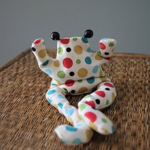 Könnte beinhalten: Ein Stofffrosch-Spielzeug mit cremefarbenem Grund und einem bunten Punktemuster in Rot, Blau, Grün und Schwarz. Das Spielzeug hat zwei schwarze, kugelförmige Augen auf dem Kopf und zwei lange, herabhängende Beine.