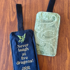 May include: Two rectangular luggage tags with black elastic loops. One tag is dark blue with an embroidered green dragon and the gold text "Never laugh at live dragons! JRR Tolkien". The second tag features a light green and white paisley pattern.