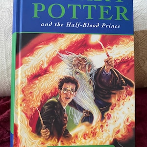 May include: A hardcover book titled Harry Potter and the Half-Blood Prince by J.K. Rowling, published by Bloomsbury. The cover features a vibrant illustration of a young boy with glasses and a wizard with a long white beard holding wands.
