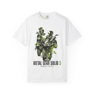 Peut inclure: Un t-shirt blanc à manches courtes avec une impression graphique représentant deux personnages dans des tons de vert et de noir. Sous l'illustration, le texte "METAL GEAR SOLID 3 SNAKE EATER" est inscrit en noir et vert sur la poitrine.