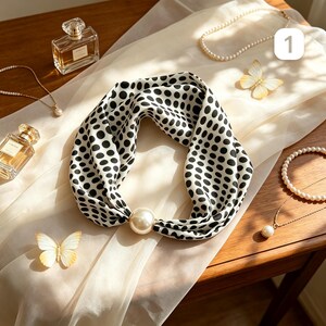 May include: A white silk scarf featuring a black polka dot pattern, secured with a large, round, cream-colored faux pearl ring. The scarf is arranged on a sheer white fabric alongside two glass perfume bottles, two pearl necklaces, and two decorative butterflies.
