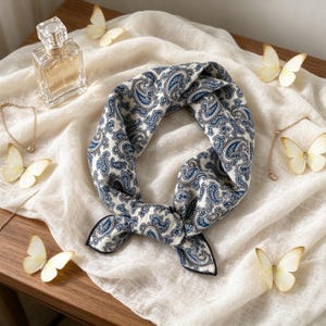 Può includere: Un foulard quadrato con motivo paisley blu scuro e bianco, annodato a cerchio. Il foulard è circondato da un flacone di profumo in vetro trasparente, due delicati braccialetti a catena color oro e cinque ornamenti a forma di farfalla bianca.