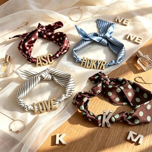 May include: Four fabric hair scrunchies with tied ribbon accents and gold or silver metal letter charms. One is maroon with white polka dots, one is light blue with stripes, one is white with black stripes, and one is brown with green polka dots.