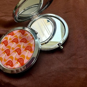 Op de afbeelding: Een ronde, zilverkleurige metalen compactspiegel die openstaat en twee reflecterende oppervlakken toont. Het buitendeksel heeft een herhalend patroon van oranje en witte paddenstoelen tegen een achtergrond van golvende roze en oranje strepen.
