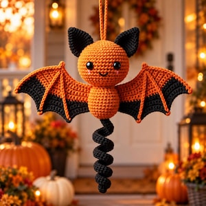 May include: A crocheted bat ornament featuring a round orange head with black ears, black eyes, and a small smile. The body is orange with wide, outstretched wings that are orange at the top and black at the bottom, ending in a long black spiral tail.