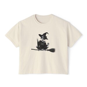 May include: A cream-colored short-sleeve t-shirt featuring a black graphic of a skeleton witch wearing a hat, sitting on a broomstick, making a peace sign, and holding a coffee cup. The skeleton wears a shirt with the text RAISING HELL & DRINKING COFFEE.