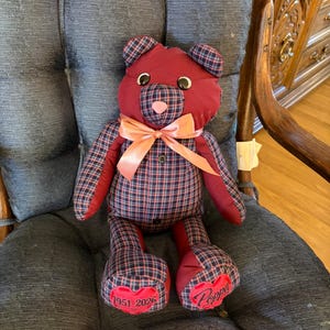 May include: A stuffed teddy bear crafted from maroon fabric and a navy, red, and white plaid pattern. It features a peach-colored satin ribbon around its neck, black button eyes, and two red heart patches on its feet embroidered with 1951-2026 and Poppa.