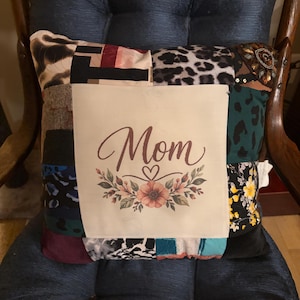 May include: A square patchwork throw pillow featuring a central cream-colored panel with the text Mom in brown script, accented by a small heart and a floral illustration. The surrounding fabric patches include leopard print, floral, and solid textures.