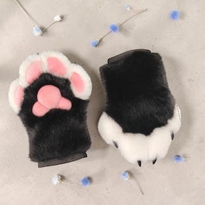 以下が含まれることがあります： 黒いフェイクファー、白いファーのアクセント、手のひら側に柔らかいピンクの肉球パッドをあしらった、動物の足の形をした指なし手袋。指先には質感のある小さな黒い爪のディテールが施されています。