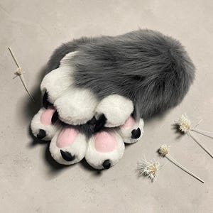 May include: A plush animal paw glove featuring long grey faux fur on the top and white faux fur on the underside. The paw includes four soft white toes with light pink circular pads and sharp black claws extending from each toe tip.