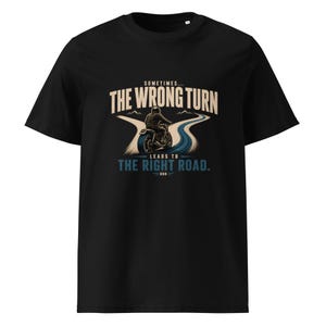 May include: A black short-sleeve t-shirt featuring a graphic of a motorcyclist on a road fork. The text reads: SOMETIMES... THE WRONG TURN LEADS TO THE RIGHT ROAD. The design uses cream, blue, and black tones to depict the rider and the winding path.